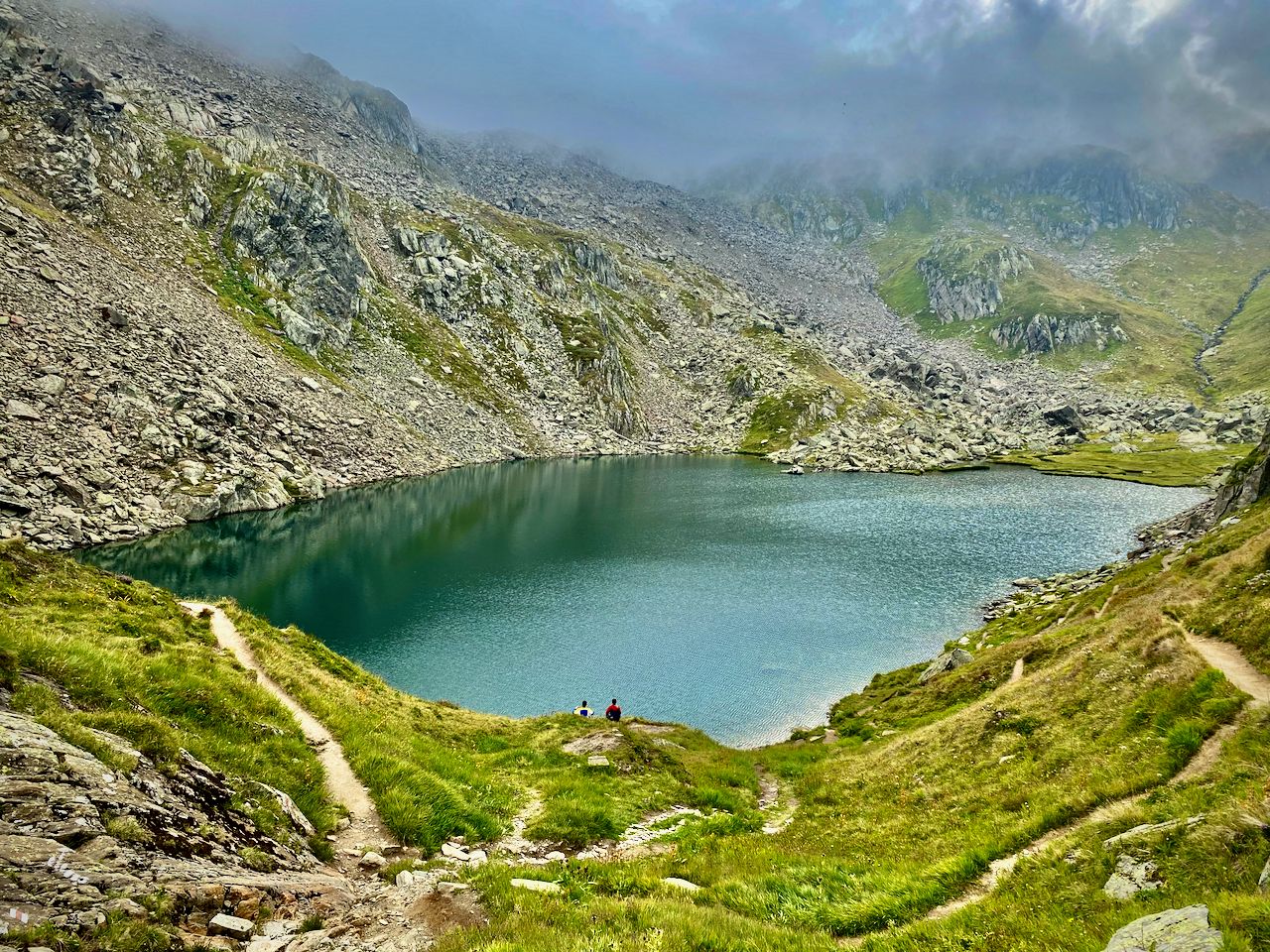 A wide angle photo of a small lake nestled between two mountains with two people sitting near the water.