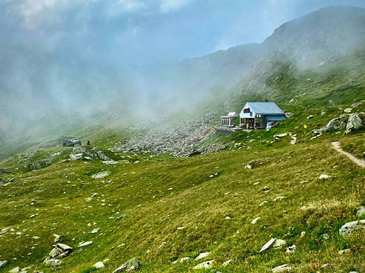 A hut nestled into the grassy side of a mountain with clouds swirling around it.