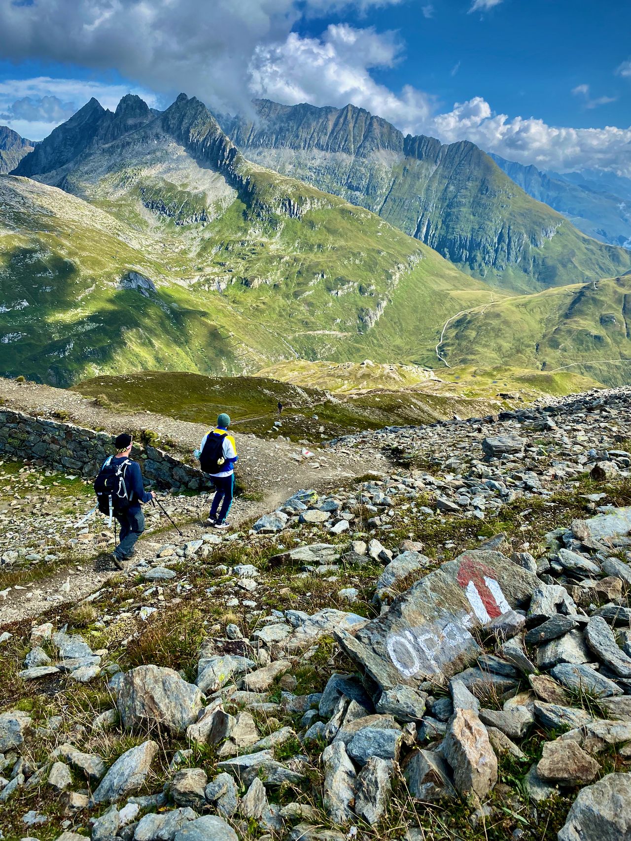 A wide angle of the mountains with two men in the foreground walking downward along a path marked O'pass.