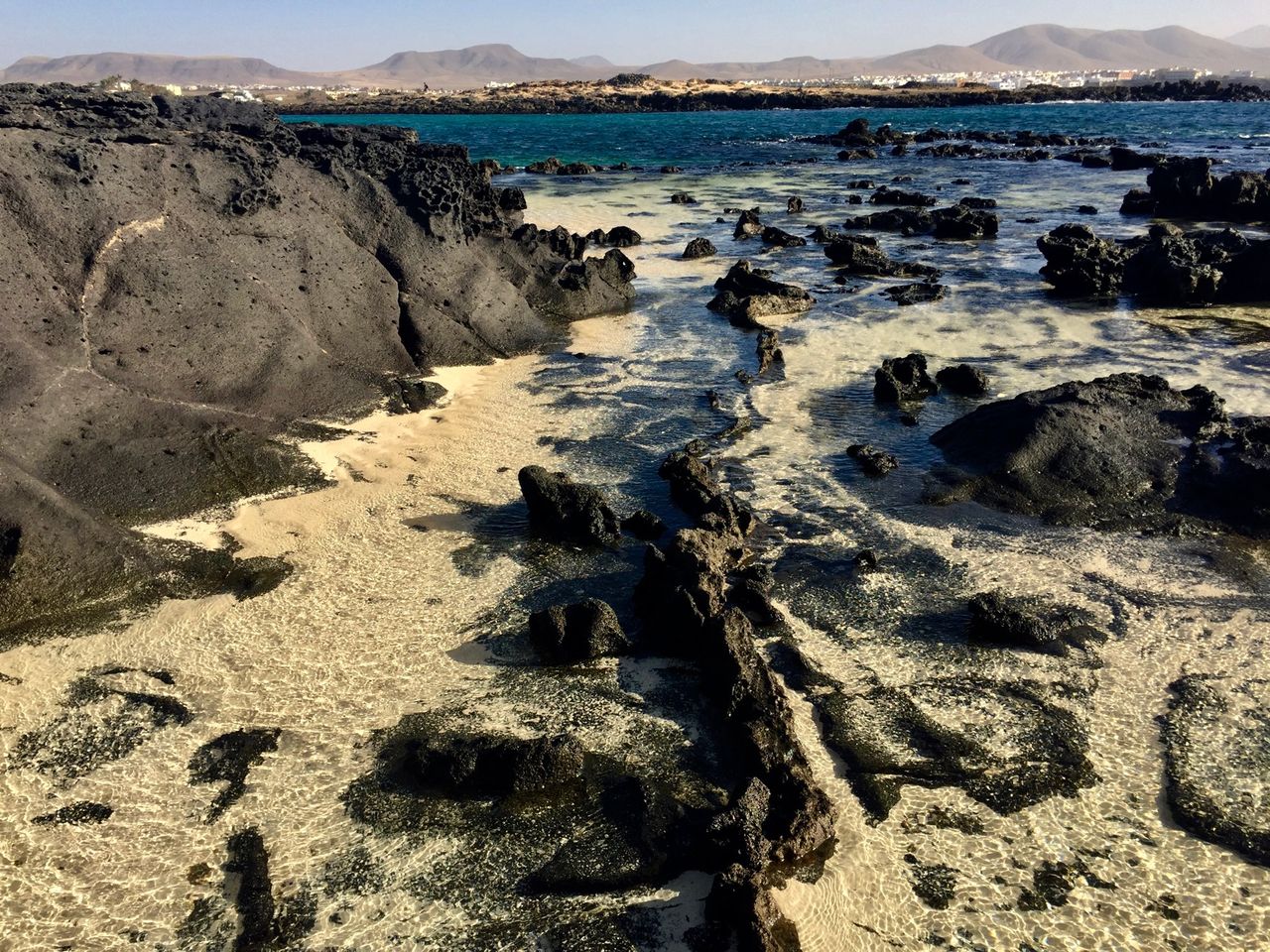 Black lava rocks in shallow water covered by sand.