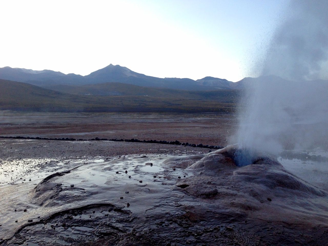 A geyser spewing steam with mountains behind it.
