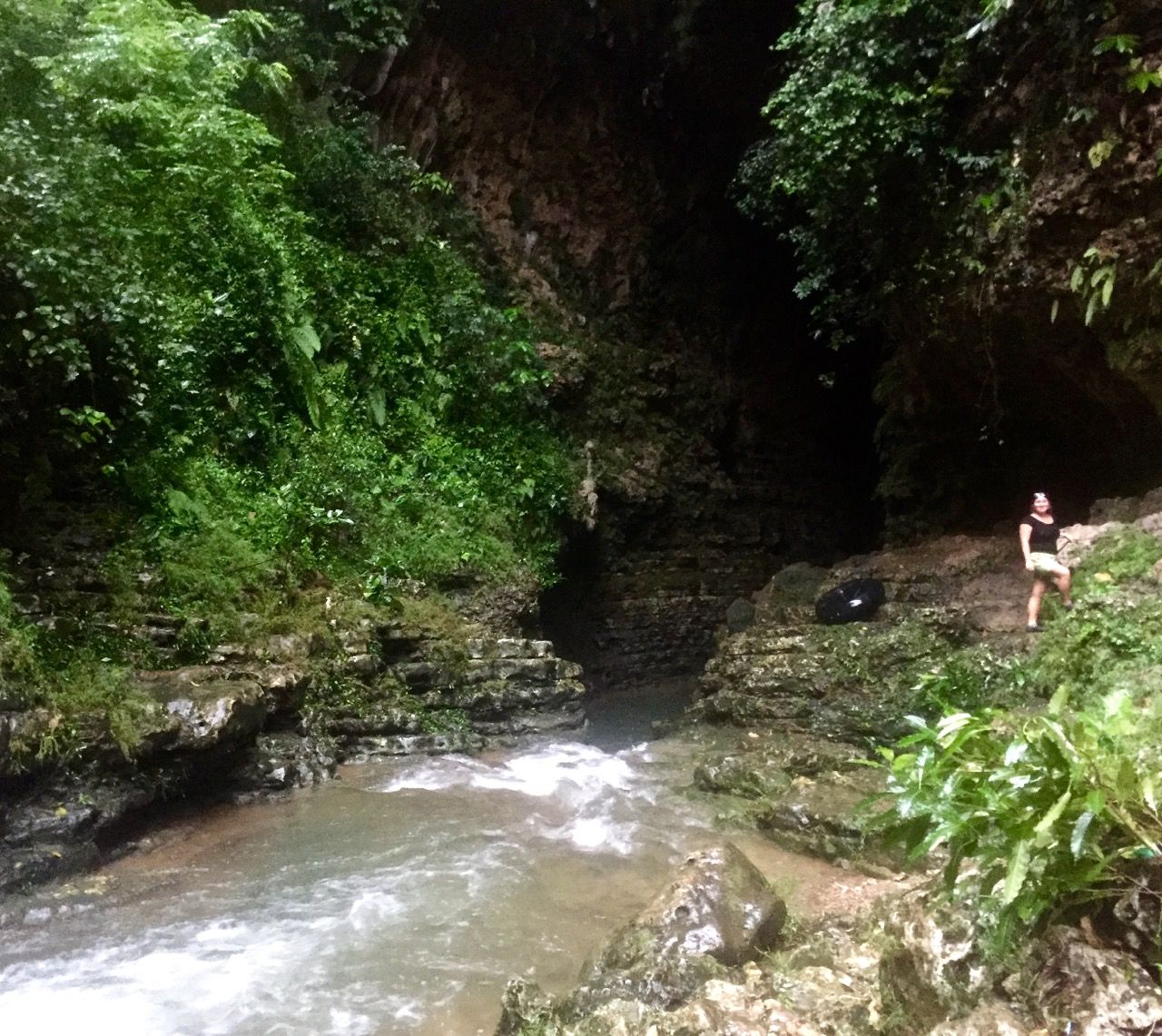 Woman posing in front of dark cave with a river flowing into it.