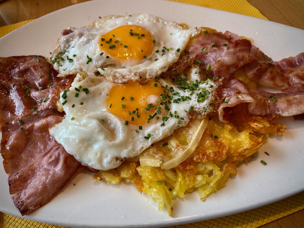 A full plate of fried shredded potatoes, fried ham, fried bacon, melted raclette, and two fried eggs with chives on top.