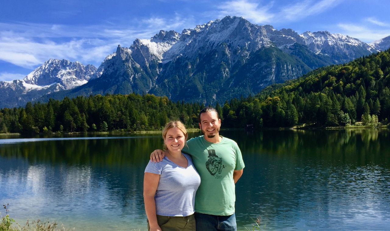 Couple smiling in front of a lake with mountains in the background.