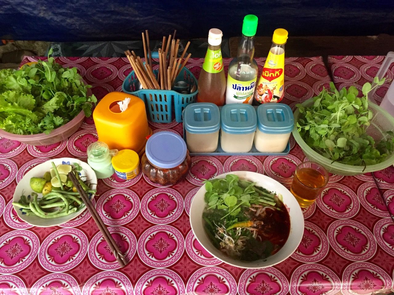 A table with fresh greens, fish sauce, chiles, and a bowl of noodle soup.