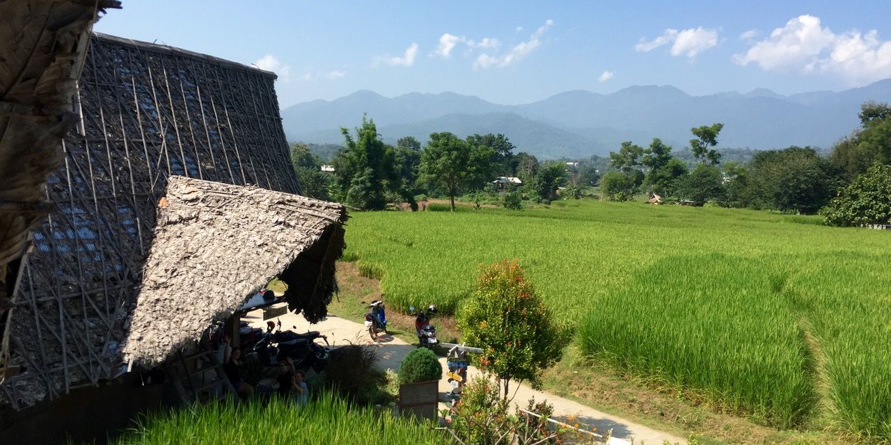 Bamboo and leaf-roofed huts set against rice fields and mountains in the background.