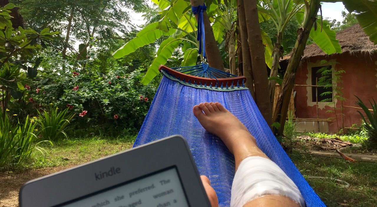 First-person view of man reading book in hammock with a bandaged leg.