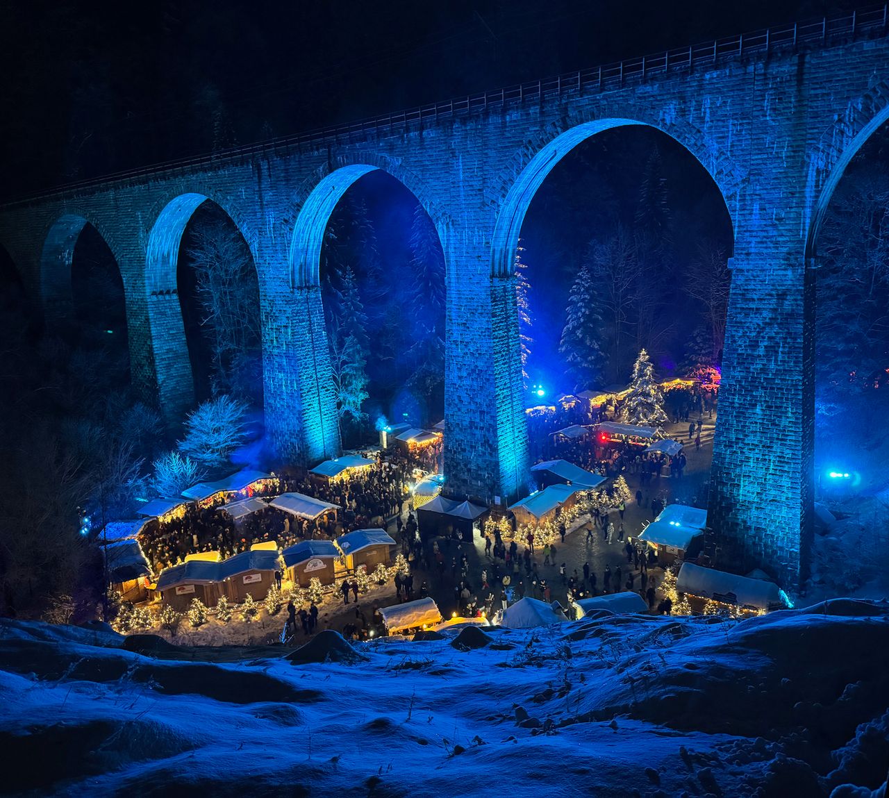 A Christmas market nestled under an old stone bridge spanning a valley, with everything lit up in blue floodlights.