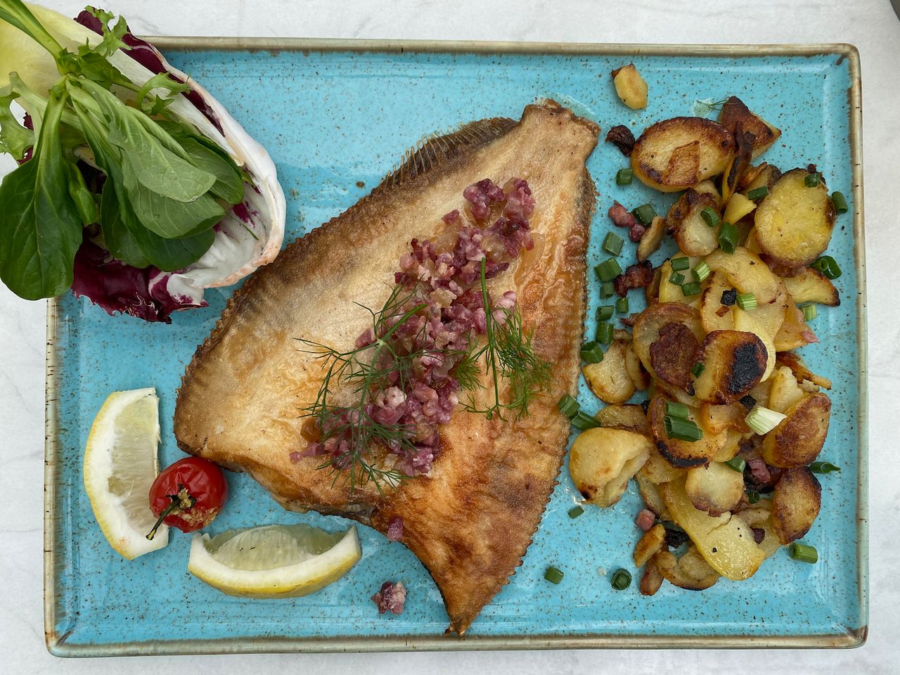 A turquoise plate with a whole fried fish, fried potatoes and some green leafy garnishing.